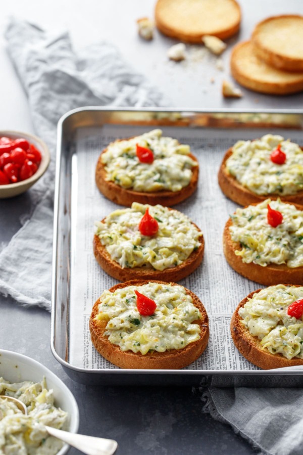 Cheesy Artichoke Crostini toasts arranged on a silver baking sheet with a bowl of artichoke topping and pepper drops on the side.