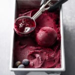 A metal loaf pan filled with Blueberry Ginger Sorbet, with an ice cream scoop and a perfect ball of sorbet.