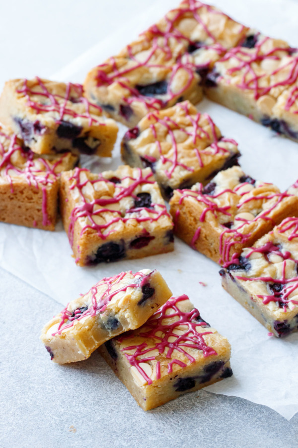 Scattered squares of Blueberry White Chocolate Blondies on parchment, one with a bite out of it to show texture.