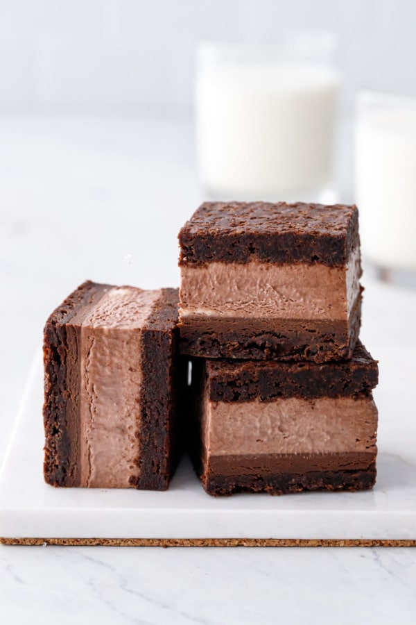 Pile of three Malted Fudge Brownie Ice Cream Sandwiches, cut into squares to show the different layers, on a marble board with glass of milk in the background.