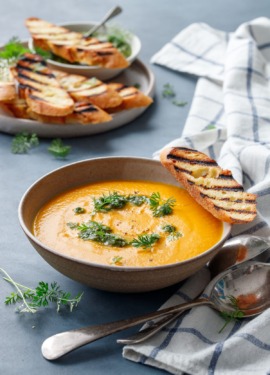Ceramic bowl with carrot soup, topped with carrot pesto, carrot greens and a slice of grilled baguette bread, on a blue background with a plate of more bread in the background.