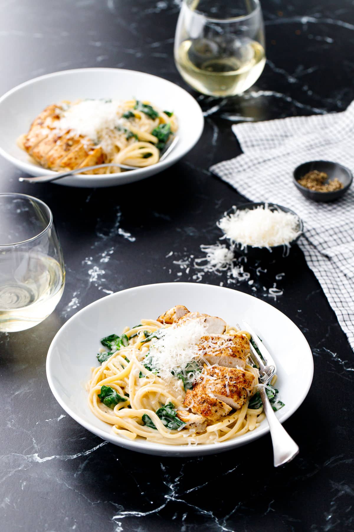 Two white bowls with Creamy Chicken Florentine Pasta, topped with a pile of shaved parmesan cheese, glasses of white wine and small bowl of cheese and black pepper on the side.