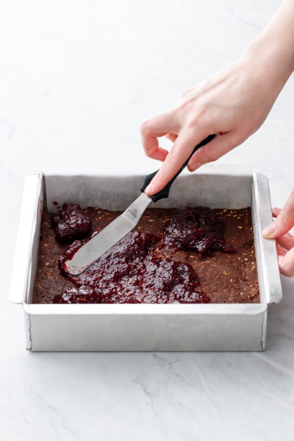 Spreading a layer of raspberry jam onto the chocolate base in an aluminum baking pan.