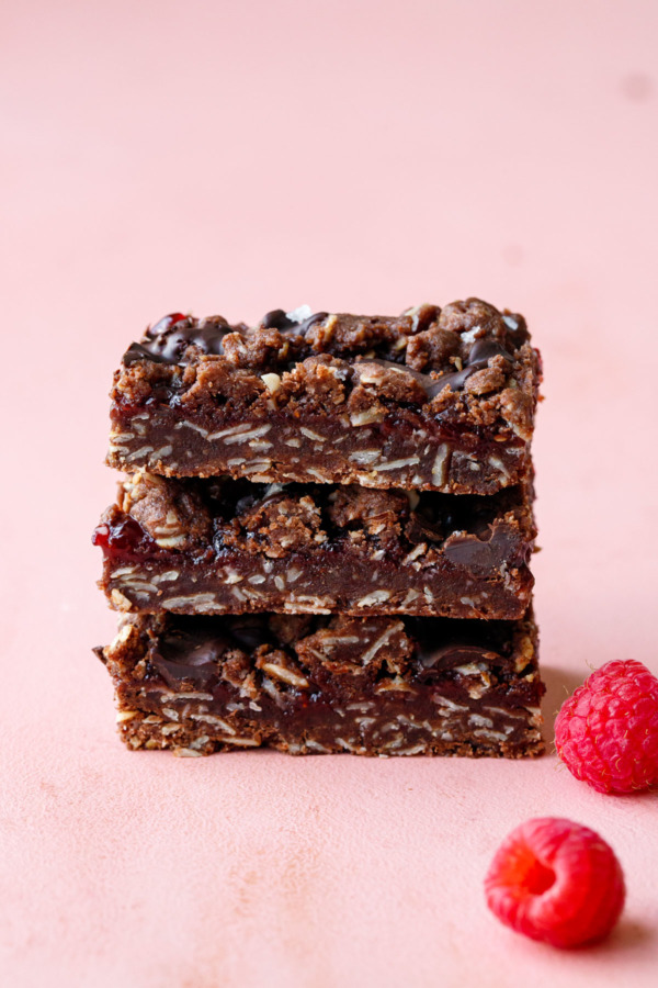 Neat stack of three rectangular-cut Chocolate Raspberry Crumb Bars on a pink background, with two fresh raspberries on the side.
