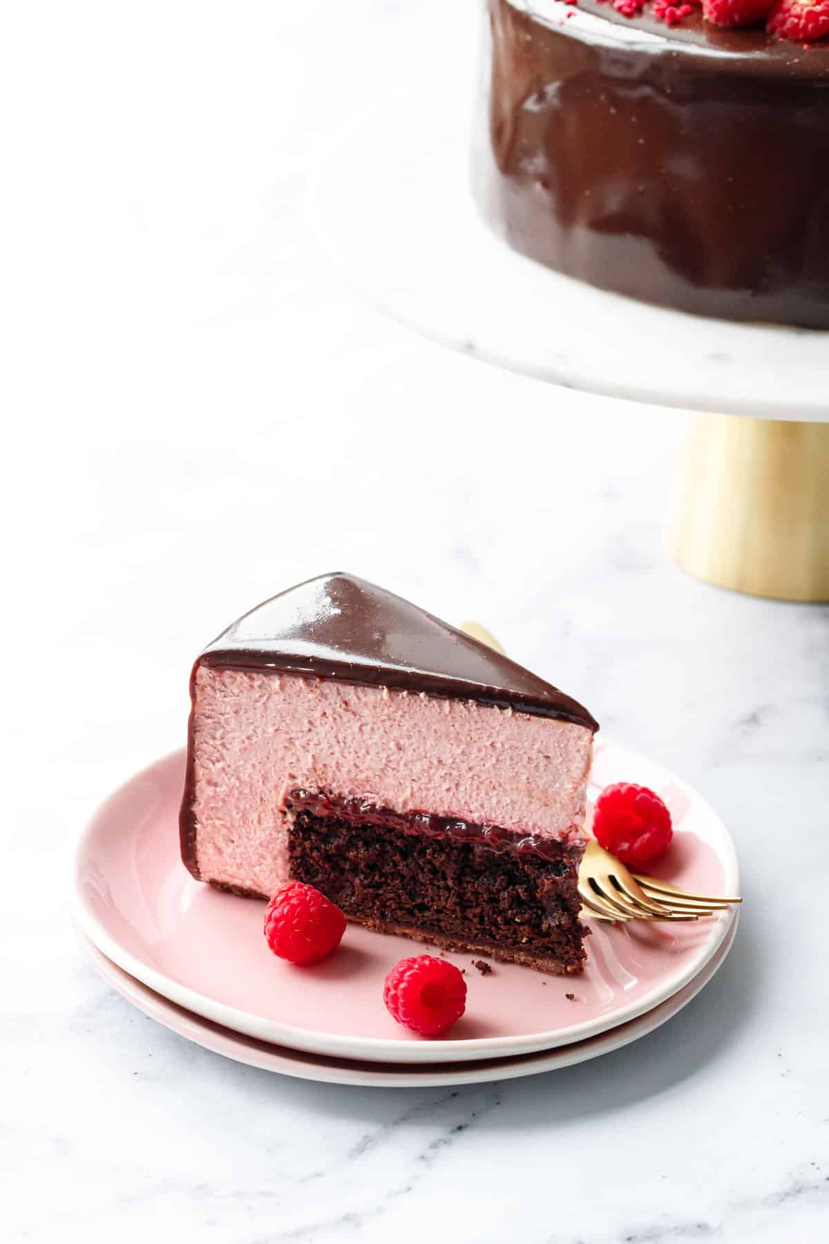 Slice of Chocolate Raspberry Mousse Cake cut into a perfect triangular slice to show the distinct layers inside (cake, jam, and pink mousse) on a pink plate with cake stand in the background.