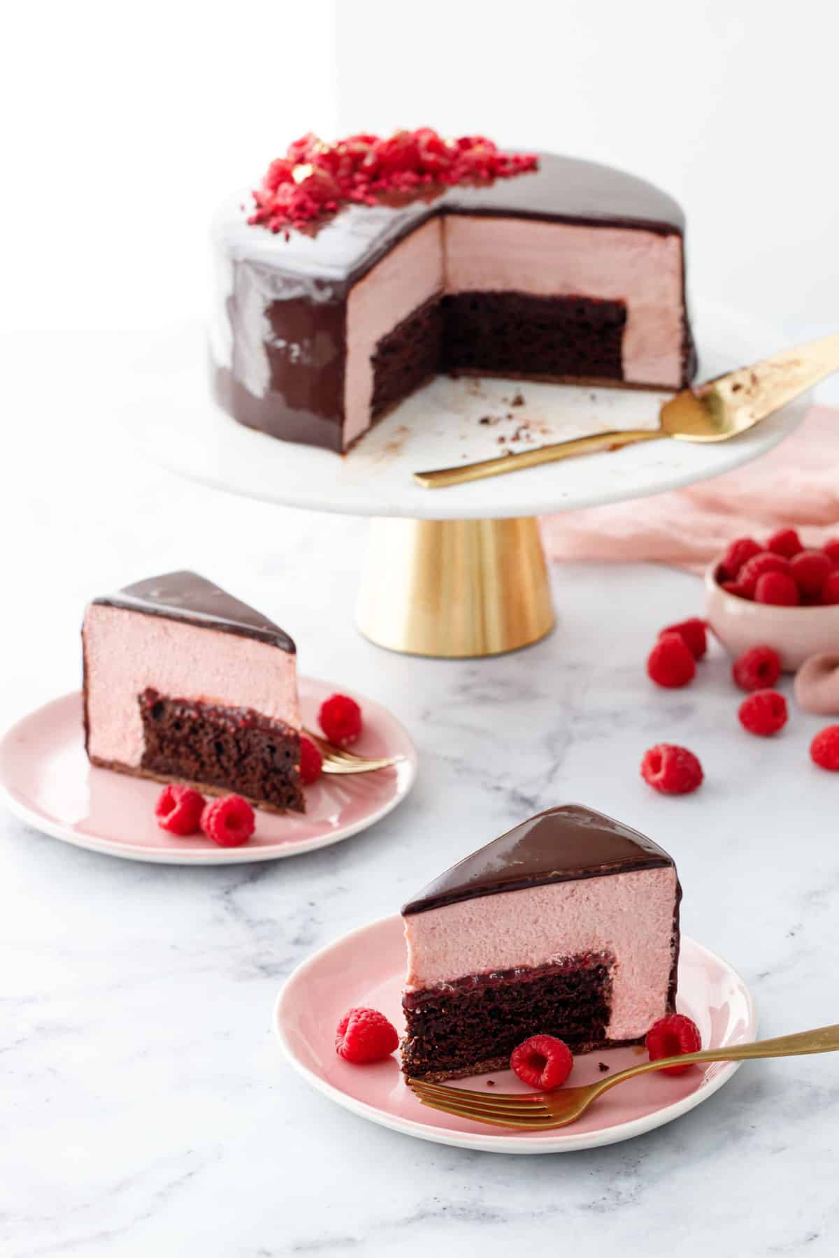 Two slices of Chocolate Raspberry Mousse Cake on pink plates, with the whole cut cake on a gold and marble cake stand in the background, with bowl of raspberries on the side.