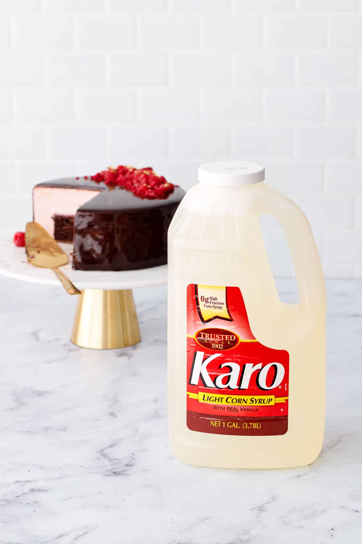 Bottle of Karo® Syrup corn syrup on white marble surface with Chocolate Raspberry Mousse Cake on a gold and marble cake stand in the background.