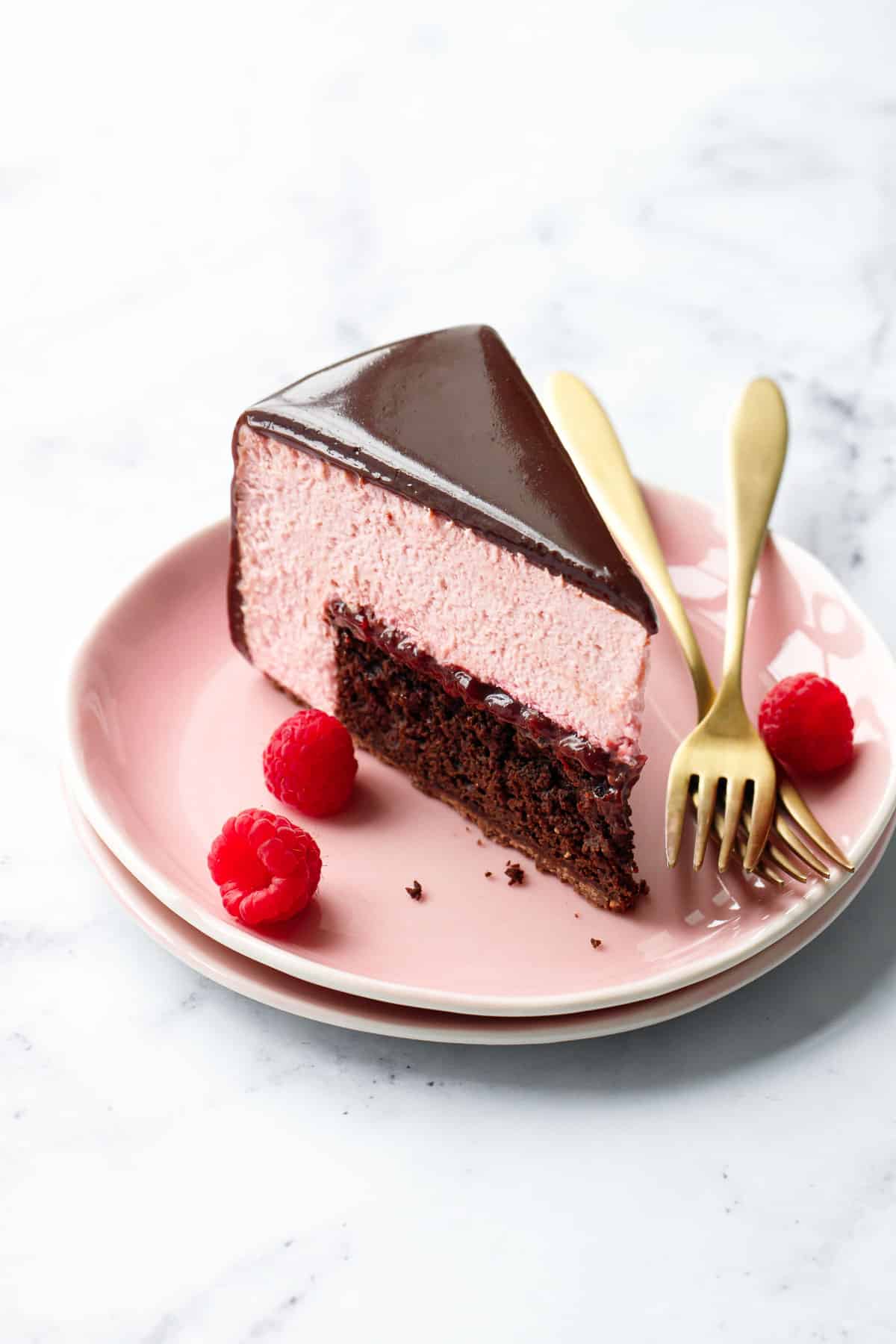 Slice of Chocolate Raspberry Mousse Cake with a super shiny chocolate glaze, on pink plates with raspberries and a gold fork.