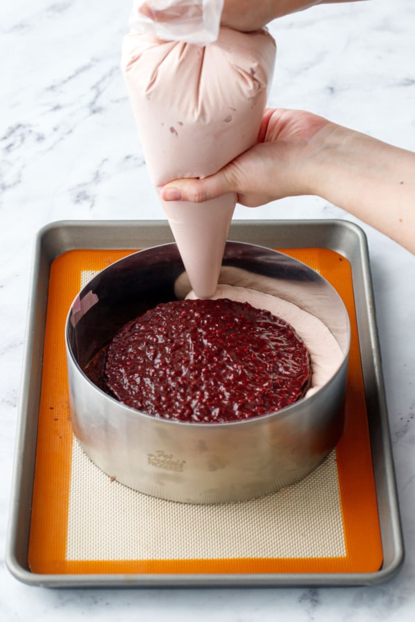 Piping the raspberry mousse around the edges of the jam-topped cake inside a cake ring.