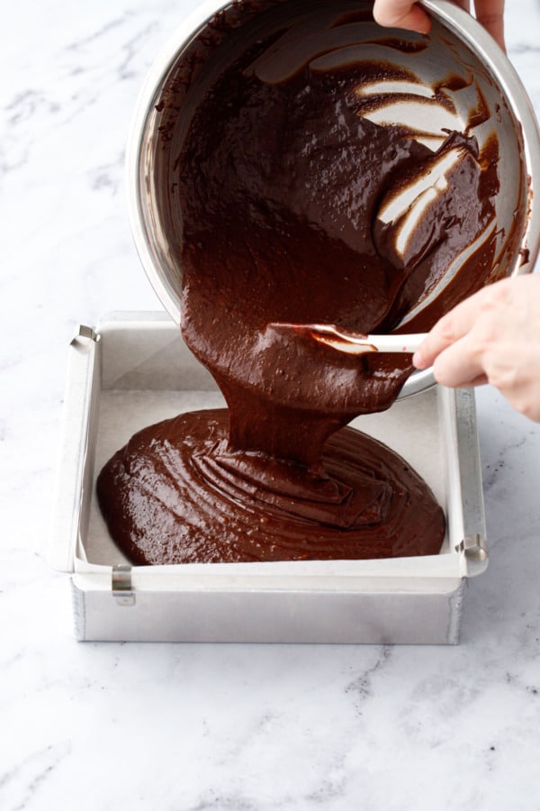 Pouring chocolate cake batter into a parchment-lined square baking pan.