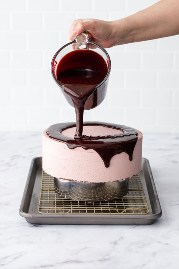Pouring dark chocolate mirror glaze on top of a light pink frozen mousse cake sitting on a wire rack on a baking sheet.