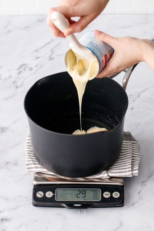Pouring sweetened condensed milk into the saucepan with the hot syrup mixture sitting on a kitchen scale.