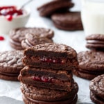 Chocolate Raspberry Sandwich Cookies, stack of cookies with one cut in half to show the fillings, with glass of milk and raspberry jam in the background.