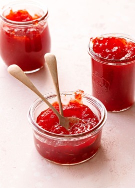 Three glass jars of Cranberry Pepper Jelly on a light pink background, one har with two spoons.