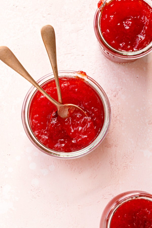 Overhead, three jars of cranberry pepper jelly with two spoons on a light pink background