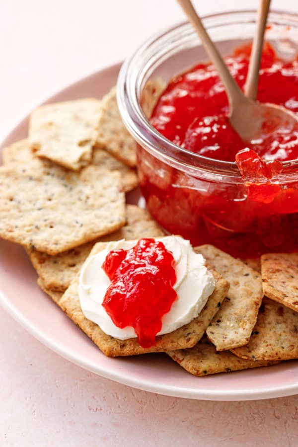 Plate of seeded crackers and a jar of cranberry pepper jelly, one cracker spread with cream cheese and a dollop of jelly