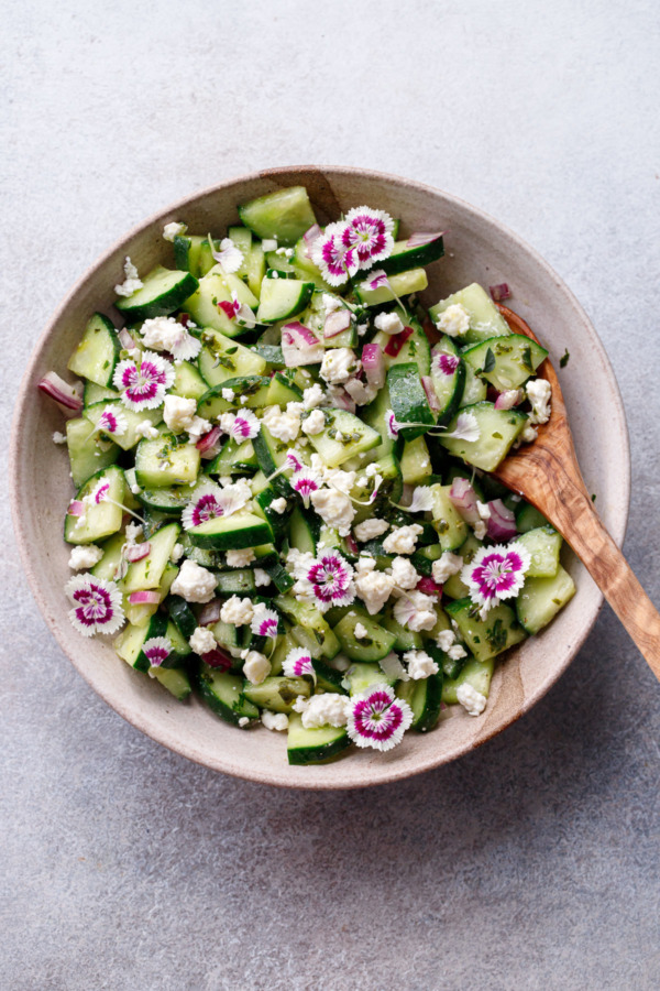 Overhead, bowl with wooden spoon and cucumber feta salad with edible flower garnish