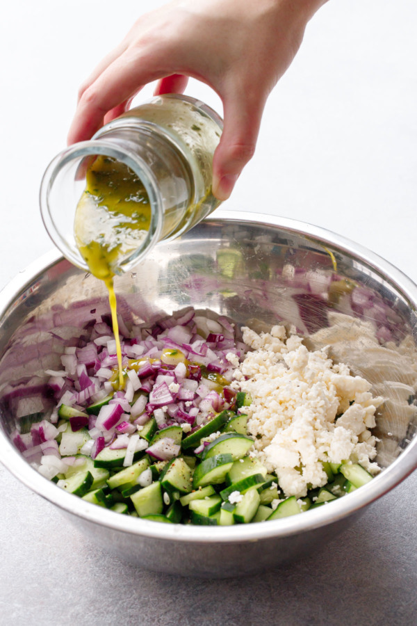 Metal prep bowl, hand pouring a glass pitcher of dressing on top of raw ingredients (cucumber, red onion, feta)