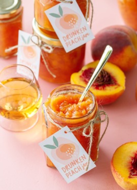 Open jar of peach jam with a gold spoon, on a pink background with more jars of jam, cut peaches and a glass of bourbon.
