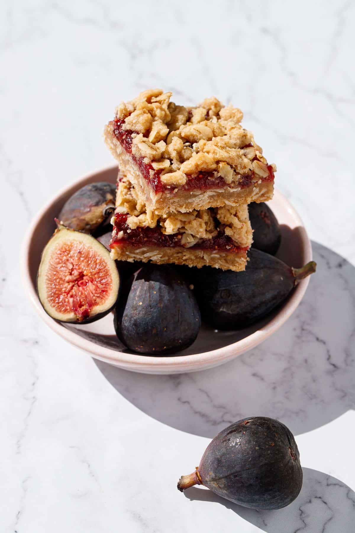 Two square Fig, Apple & Vanilla Crumb Bars sitting on top of a bowl of fresh mission figs, one cut in half to show the vibrant inside.