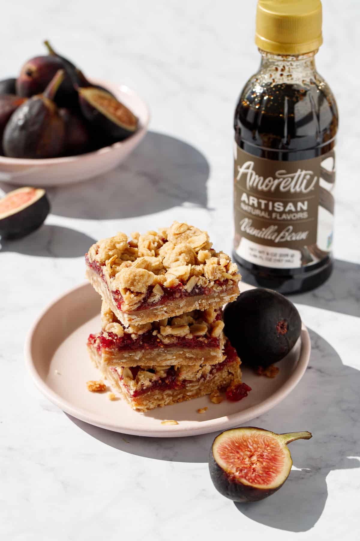 Plate with stack of three Fig, Apple & Vanilla Crumb Bars, with a bowl of fresh figs and one cut fig in the foreground, bottle of Amoretti Vanilla Artisan flavor in the background.