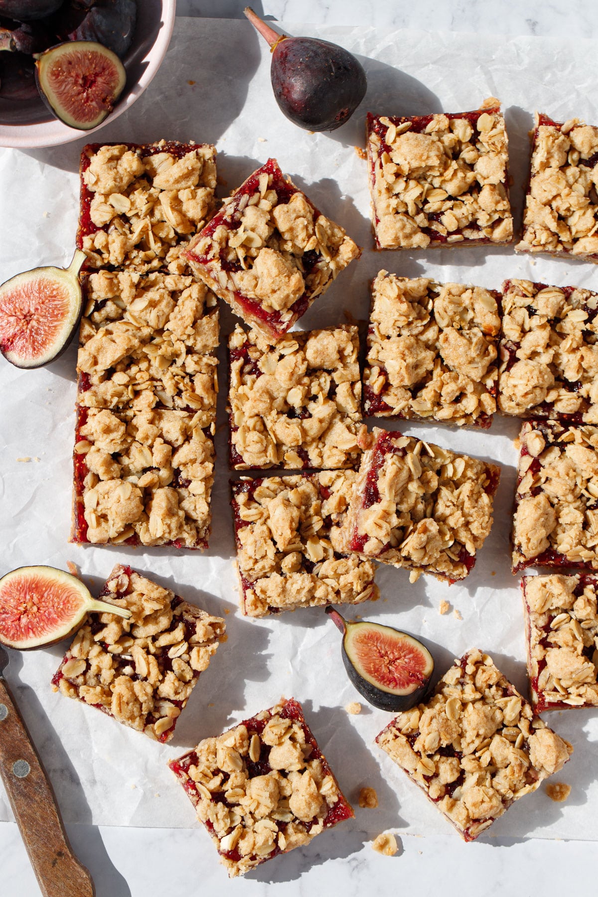 Overhead, messy arrangement Fig, Apple & Vanilla Crumb Squares on a marble background with fresh figs, a few cut in half, and a knife on the side.