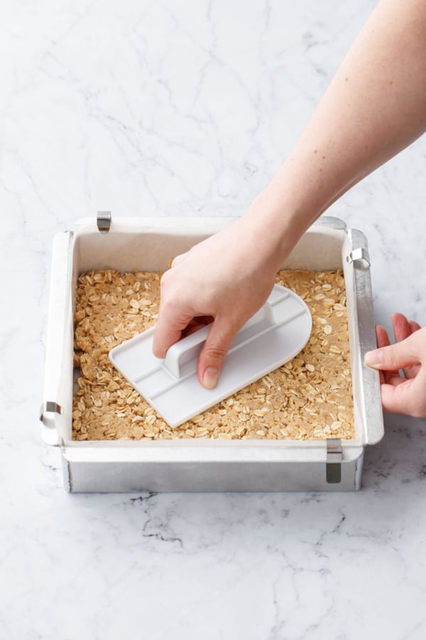 Using a fondant smoother to press the oat mixture into the parchment lined baking pan.
