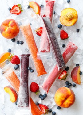Overhead, Freezer Jam Ice Pops in three different color/flavors, scattered on crushed ice cubes with fresh strawberries, blueberries, and peaches scattered around.