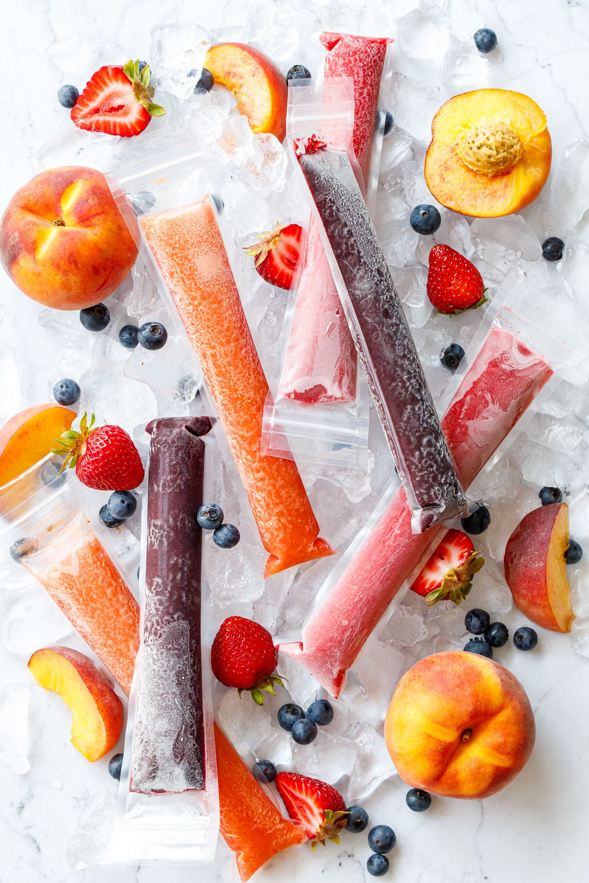 Overhead, Freezer Jam Ice Pops in three different color/flavors, scattered on crushed ice cubes with fresh strawberries, blueberries, and peaches scattered around.