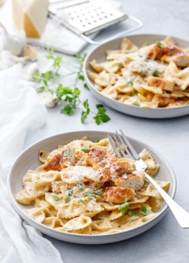 Two shallow bowls with Creamy Garlic Chicken Pasta, napkin and forks and a piece of parmesan cheese in the background.