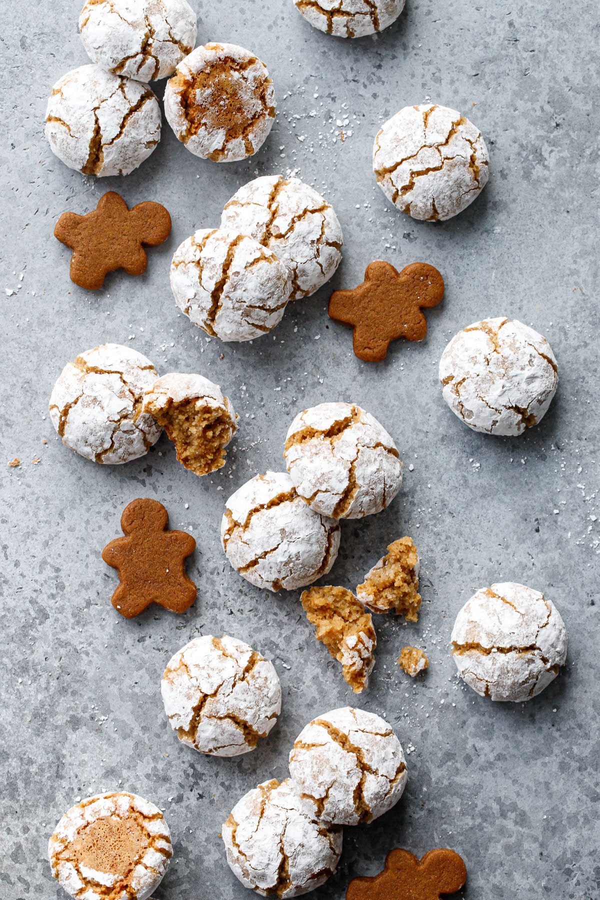 Overhead, scattered Gingerbread Amaretti Cookies on a gray background, some cookies broken and crumbled and a few mini gingerbread men interspersed among the other cookies.