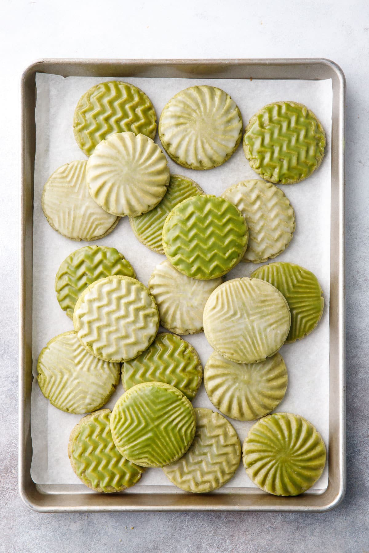 Overhead, messy arrangement of Glazed Matcha Sugar Cookies on a parchment lined baking sheet; cookies have white or green glaze and geometric stamped designs.