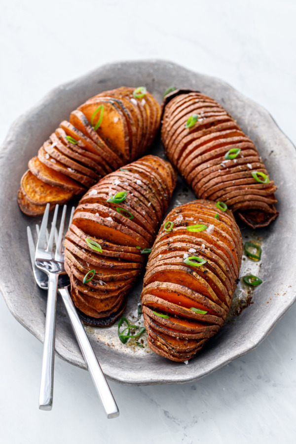 Four Spiced Hasselback Sweet Potatoes arranged on a vintage metal serving plate with two forks, on a white marble background.