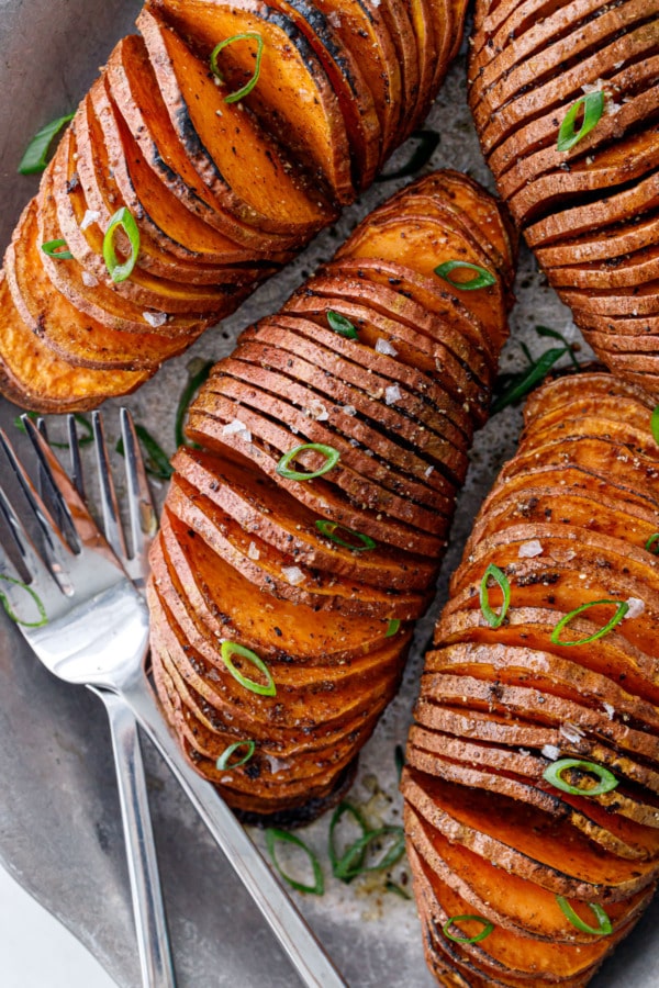Closeup overhead of four Spiced Hasselback Sweet Potatoes on a metal plate with forks, topped with green onions.