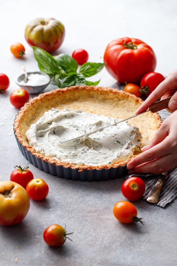 Spreading basil goat cheese into a pre-baked tart shell, surrounded by heirloom tomatoes