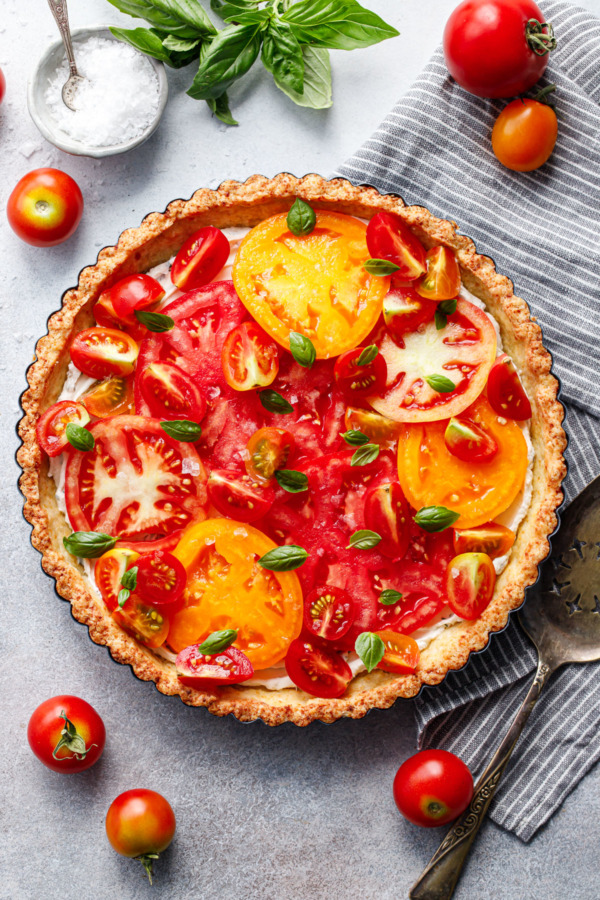 Overhead shot of heirloom tomato tart on a gray background, with tomatoes scattered around and a small bowl of flaky sea salt