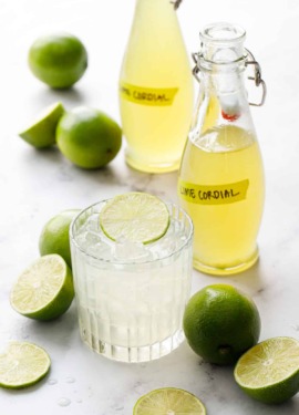 Bottles of Homemade Lime Cordial with fresh limes and a gimlet cocktail garnished with a lime slice.