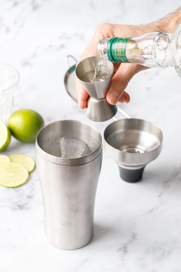 Pouring London dry gin into a jigger, then into a cocktail shaker to make a Gimlet.