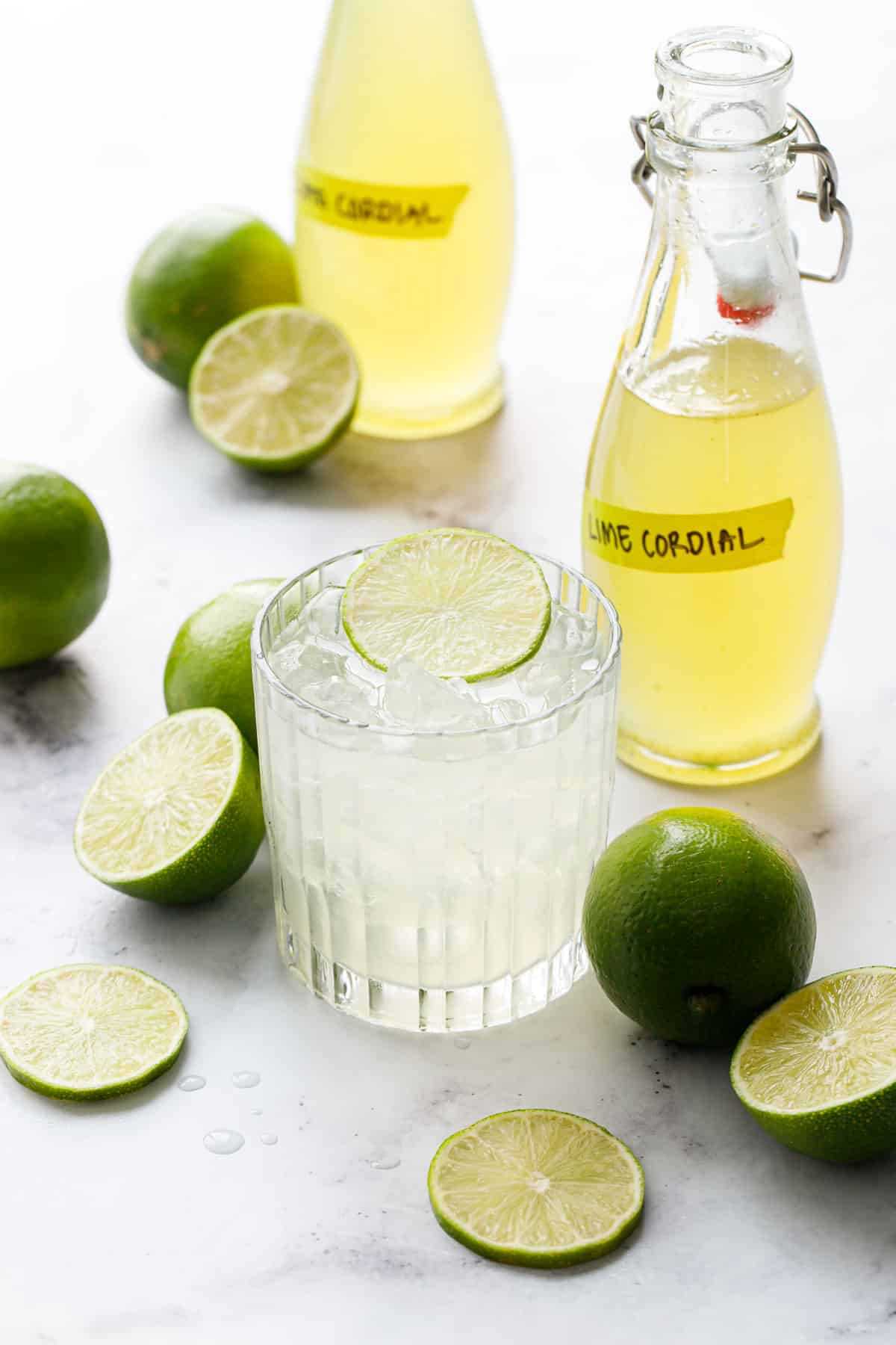 Lime Gimlet cocktail in an ice-filled lowball glass, with bottles of Homemade Lime Cordial and fresh limes scattered around.