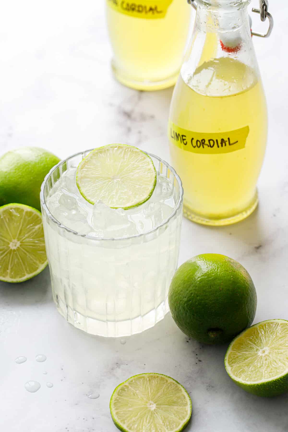 Gimlet cocktail in a lowball glass garnished with a lime slice, with two bottles of Homemade Lime Cordial and fresh limes in the background.