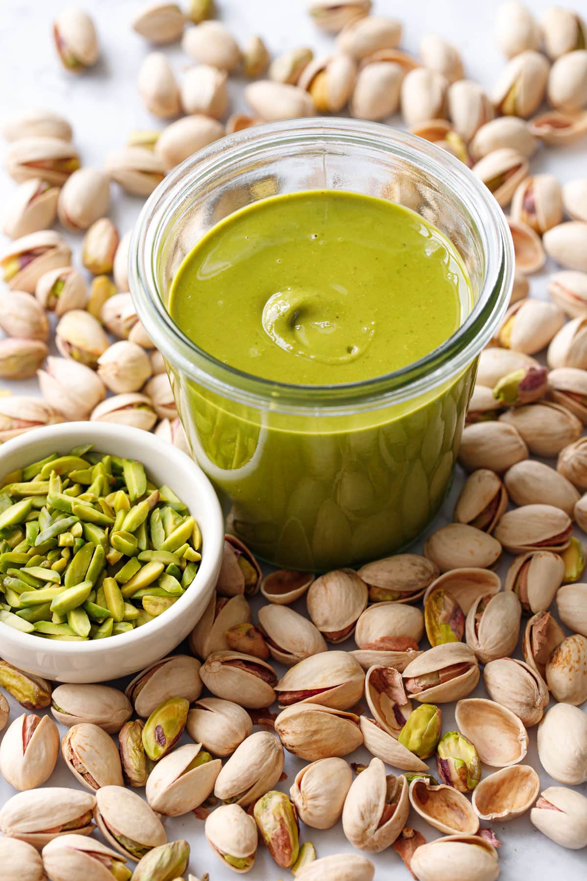 Glass jar of bright green Homemade Pistachio Butter with a bowl of green slivered pistachios, and whole in-shell pistachios scattered around.