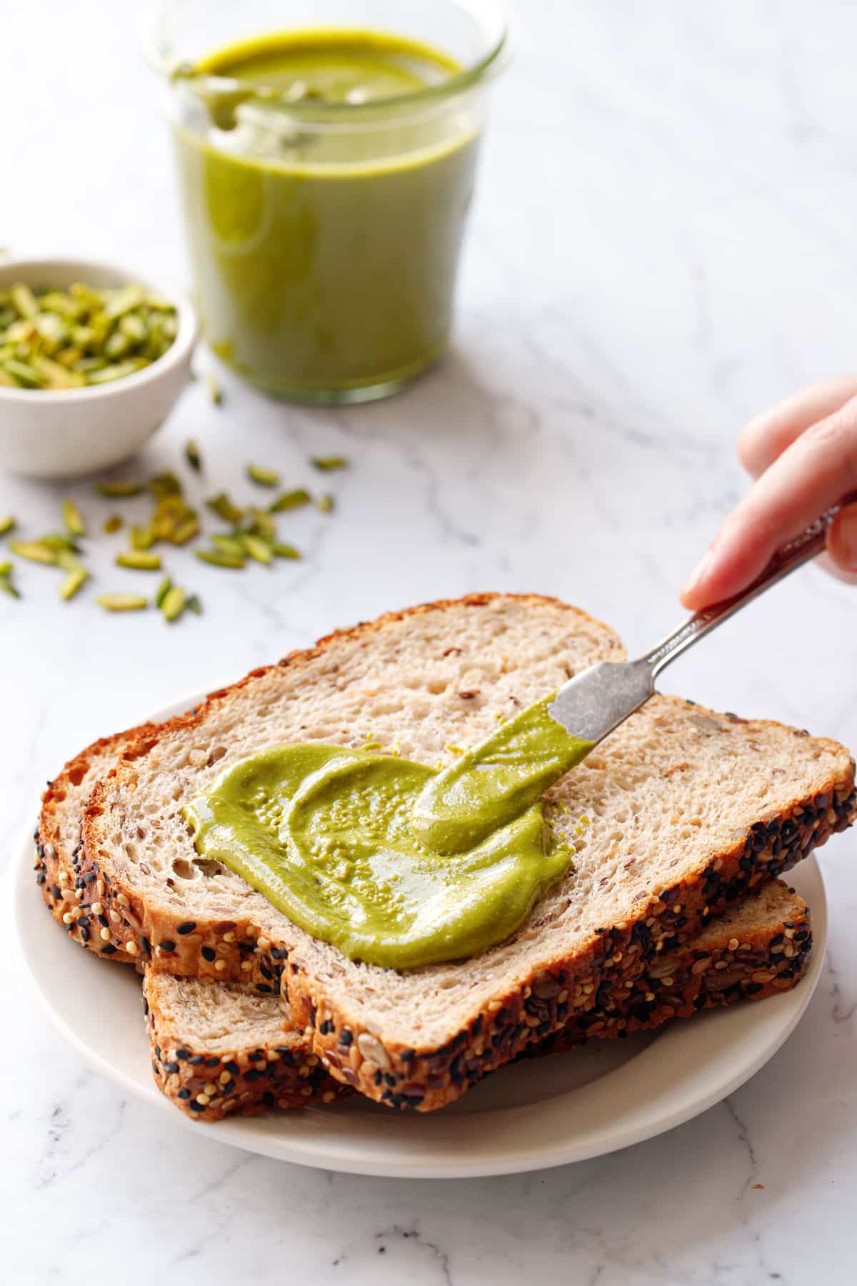Spreading bright green Homemade Pistachio Butter onto a piece of whole wheat bread, with a jar of butter and bowl of pistachios in the background.