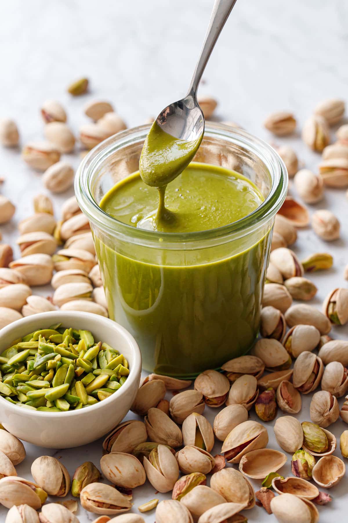 Glass jar with bright green Homemade Pistachio Butter, a spoonful lifting to show the creamy texture, with slivered and shell pistachios scattered around.
