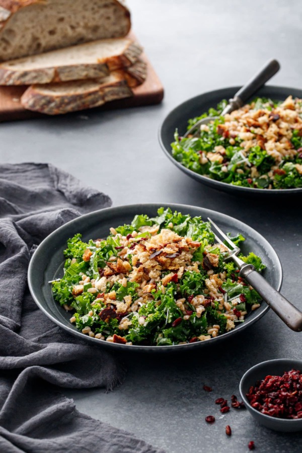 Two dark gray bowls of Kale & Farro Salad with Sourdough Breadcrumbs on a dark gray background, loaf of sourdough bread in the background.