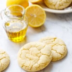 Crackly top Lemon Olive Oil Sugar Cookies on marble with a small glass pourer of olive oil, lemons, and a plate of more cookies in the background.