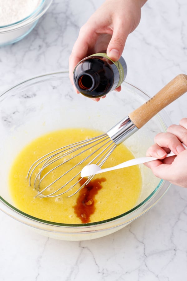 Teaspoon of vanilla extract being poured into mixing bowl with whisk.