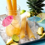 Aqua blue background with metal tray filled with ice, and mai tai cocktail, bright orange ice pops, and pineapple pieces