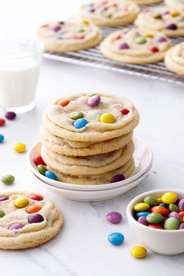 Stack of four M&M Sugar Cookies on a small cookie plate, with rack of cookies, glass of milk and bowl of M&Ms candies.