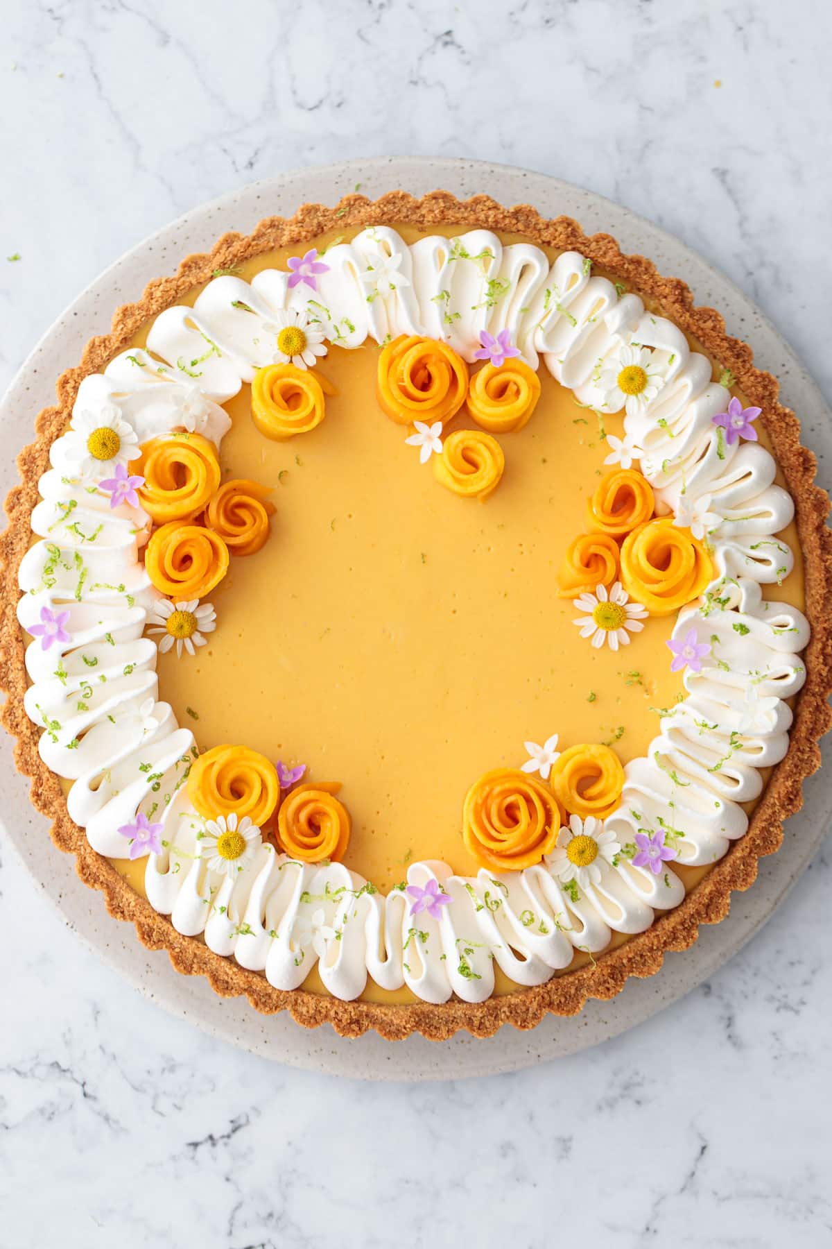 Overhead shot of a Mango Lime Tart on a marble background, decorated with a squiggle of whipped cream, mango flowers, and edible flowers.