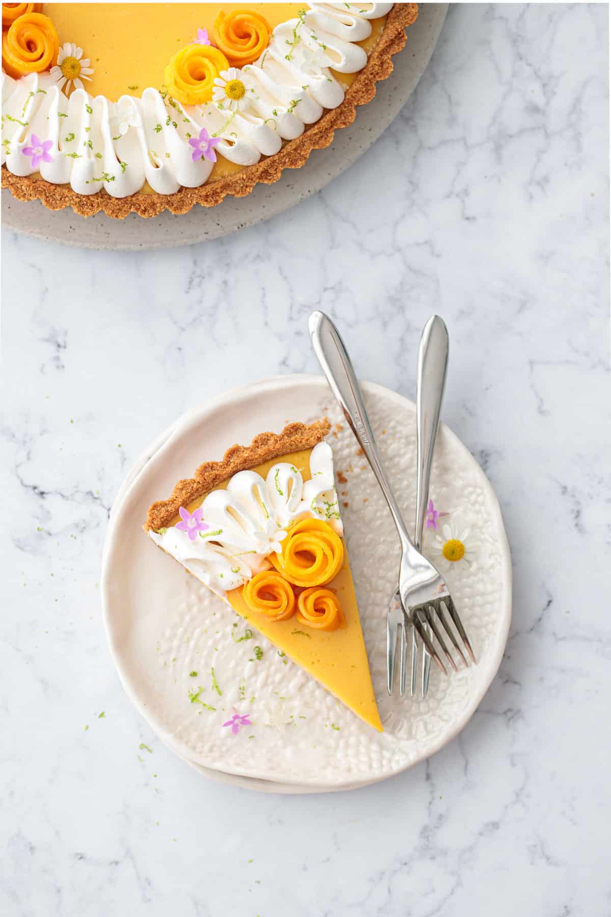 Overhead, white plate with a slice of Mango Lime Tart and two forks, topped with mango flowers and edible flowers.